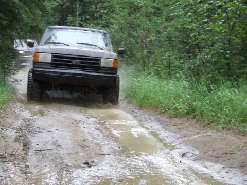 1988 Ford Bronco - A dark gray 1987 Ford Bronco navigates a muddy trail, kicking up water as it goes.