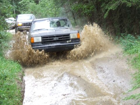 1988 Ford Bronco - Two SUVs navigate a muddy trail, kicking up large plumes of water and dirt.