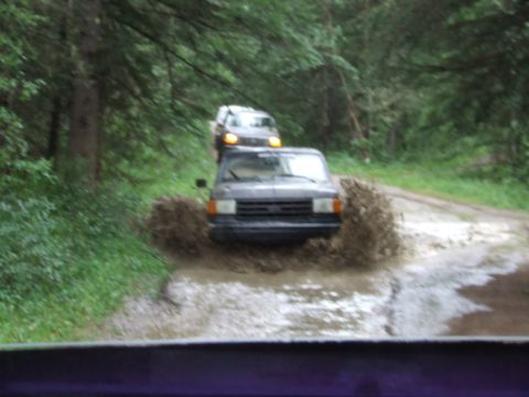 1988 Ford Bronco - Two SUVs navigate a muddy obstacle on a wooded dirt road.