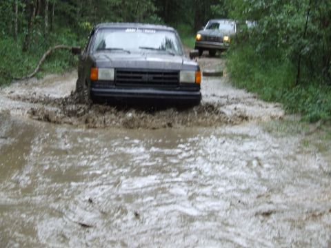 1988 Ford Bronco - Two trucks navigate a muddy trail, with the lead vehicle kicking up water and dirt.