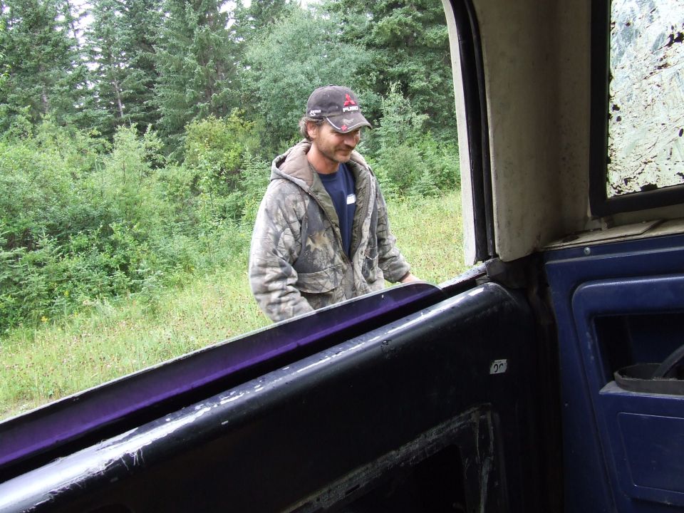 man in camo jacket and hat standing by open vehicle door, trees and grass in background