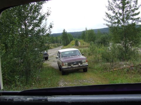 1988 Ford Bronco - Two Ford Broncos drive down a dirt road surrounded by trees and brush under an overcast sky.