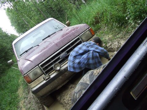 1988 Ford Bronco - A maroon Ford Bronco is stuck in mud with a person trying to help it.