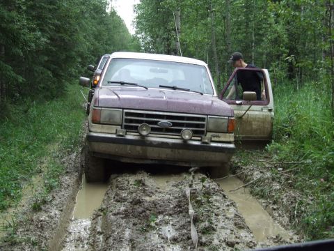 1988 Ford Bronco - Two vehicles navigate a muddy path through a forest, with one appearing to be stuck.
