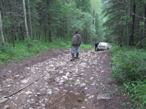 1988 Ford Bronco - A white SUV is stuck on a rough, rocky road in a wooded area, with two individuals attending to it.