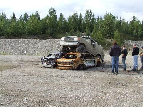 1988 Ford Bronco - A tan Ford Bronco is stacked on top of two heavily damaged cars in a gravel lot.