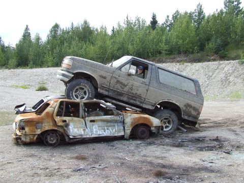 1988 Ford Bronco - A Ford Bronco is driven on top of a heavily damaged and burnt sedan, with trees and a gravel pile in the background.