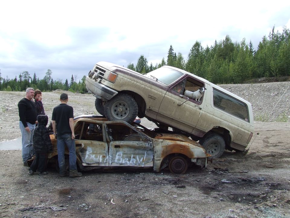 1988 Ford Bronco on top of a burnt car with 'Burn Baby!' written on its side. People and a dog are visible