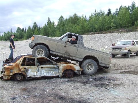 1988 Ford Bronco - A man drives a muddy Ford Bronco over a destroyed sedan, with another truck visible in the background.