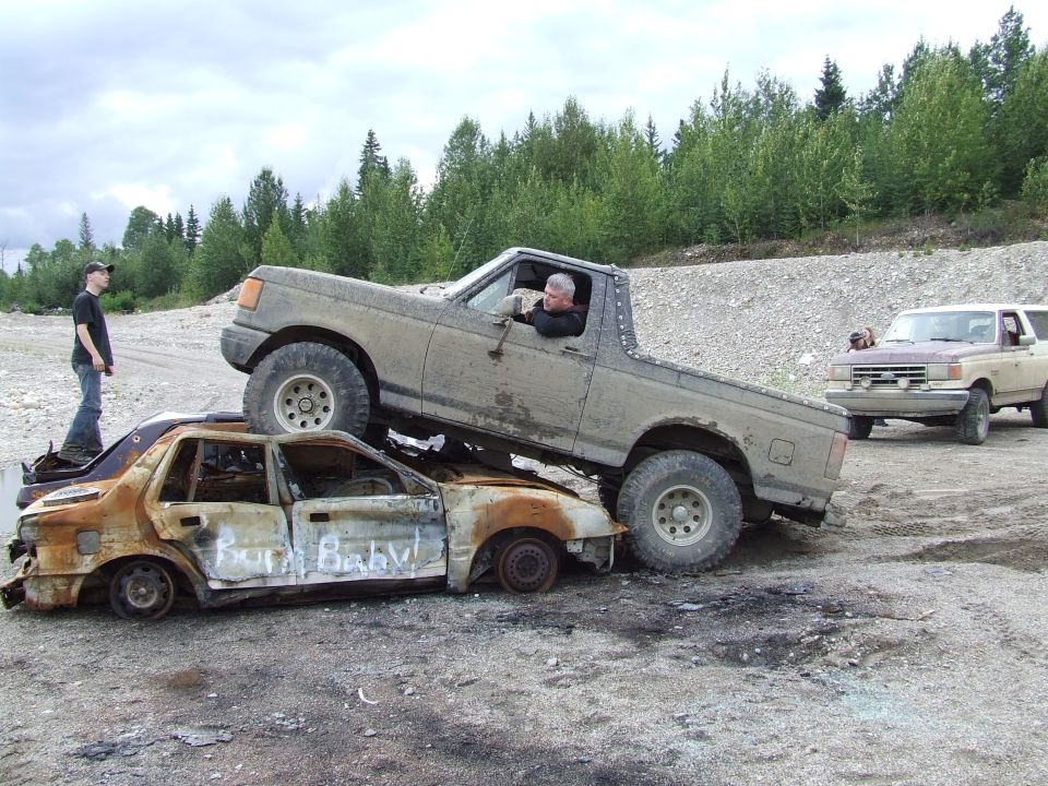 Mud-covered Ford Bronco on top of a burnt sedan, text "Born Baby!" on sedan side