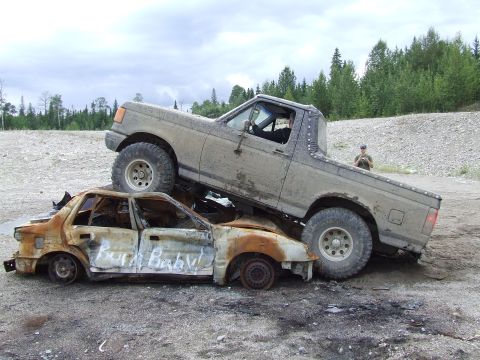 1988 Ford Bronco - A 1988 Ford Bronco is positioned on top of a heavily damaged and burnt-out sedan that has "Burn Baby!" spray-painted on its side.