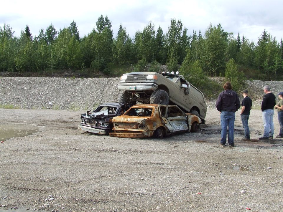 Tan off-road vehicle on top of two wrecked cars, with people watching