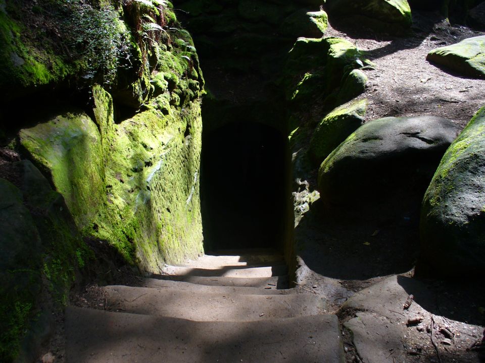 Mossy rocks form a narrow passage leading to a dark opening. Steps lead down into the passage