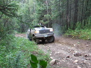 1988 Ford Bronco - Small distant photo of a lifted light-colored full-size Ford Bronco climbing a rocky, rutted dirt trail through dense spruce forest, seen h…