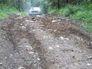 1988 Ford Bronco - Vehicle navigating a challenging, rocky path in a forest setting. SuperMotors branding visible.