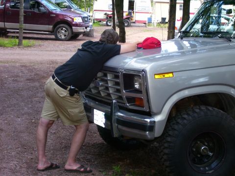 1984 Ford Bronco - Man rests his head on the grille of a silver Ford truck, with another truck behind.