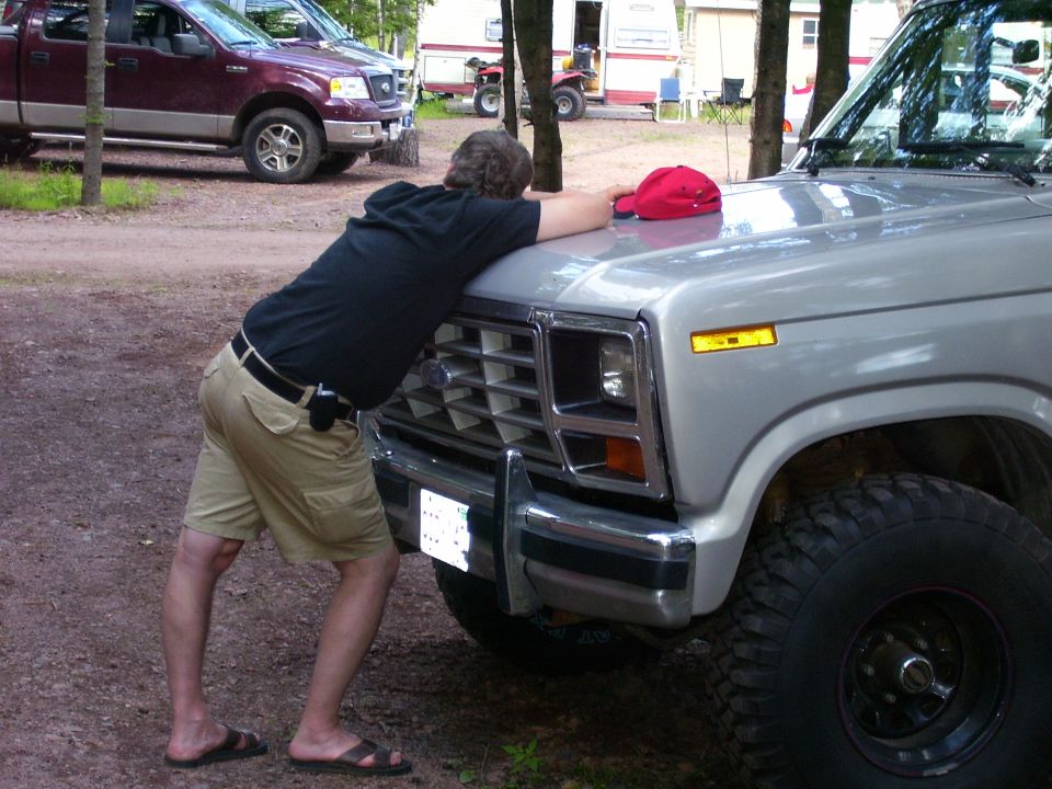 Man leaning on the grille of a silver 1980s Ford F-Series truck. A maroon Ford F-150 is parked behind it