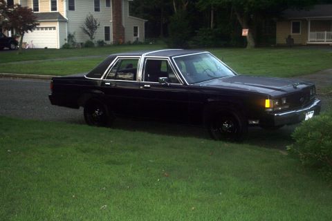 1989 Ford Crown Victoria LTD - A dark-colored sedan sits parked on a grassy lawn in front of a suburban home.