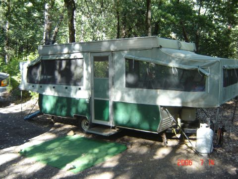 1993 Ford Bronco - A pop-up camper trailer is set up in a campsite with trees in the background. A propane tank is visible on the side.