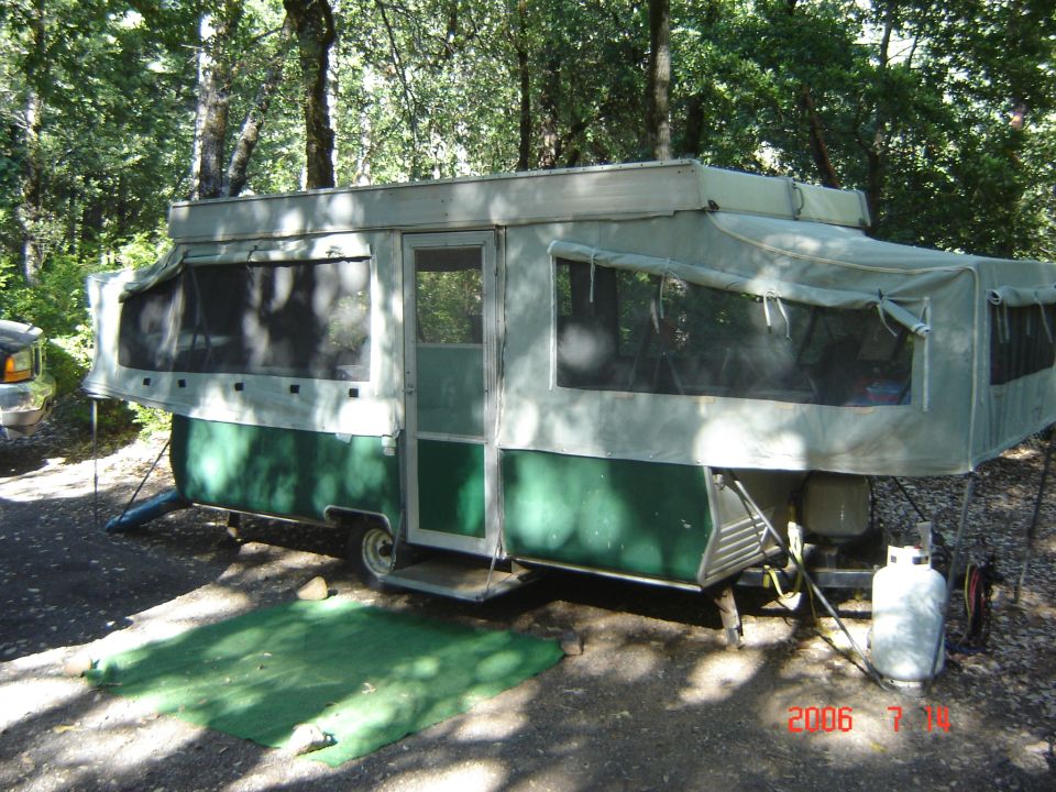 Pop-up camper trailer set up in a wooded area. Propane tank visible next to it