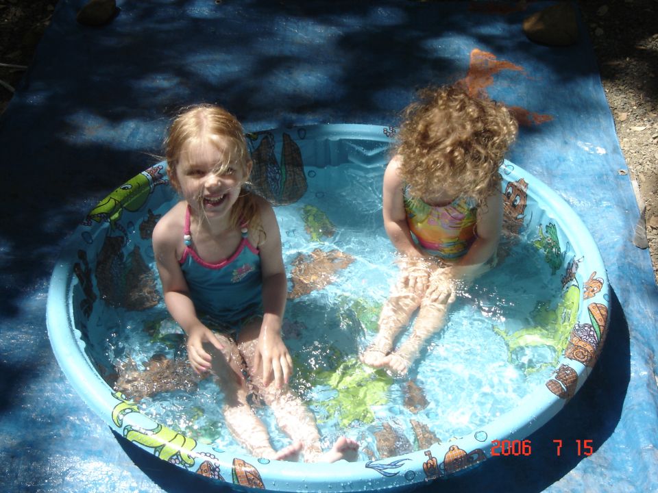 Two young girls splash in a blue kiddie pool with cartoon sea creatures on the side