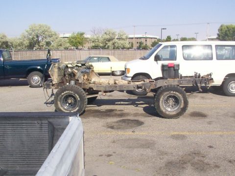 1978 Ford Bronco - A disassembled vehicle chassis sits on pavement, with other vehicles and buildings in the background.