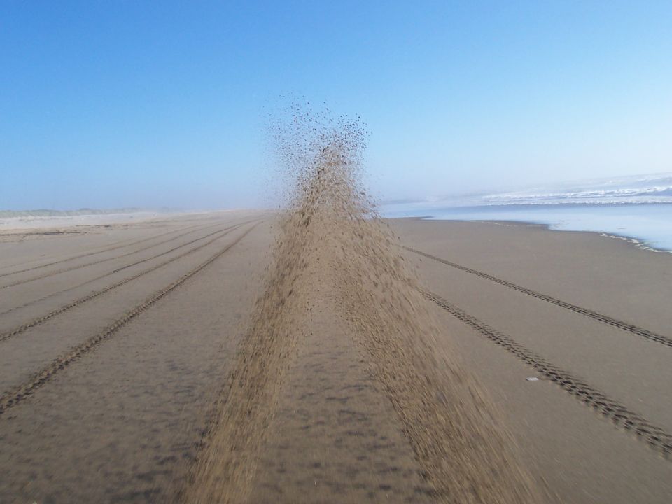 Tire tracks in the sand on a beach with the ocean in the background. Sand is being kicked up in the air