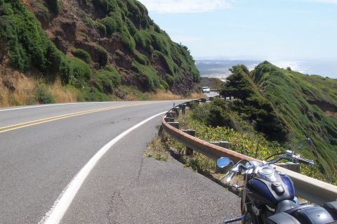 1994 Ford F-150 - A motorcycle is parked on the side of a winding coastal road, with a white car visible further down the highway.