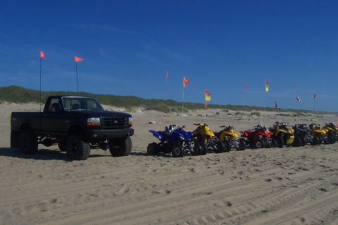 1994 Ford F-150 - A lifted black Ford F-150 sits on the beach next to a row of ATVs, with dunes and flags in the background.