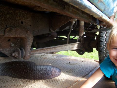 1996 Ford Bronco - A young child peeks into the frame next to a heavily rusted vehicle's undercarriage, showing an oil leak on a metal surface.