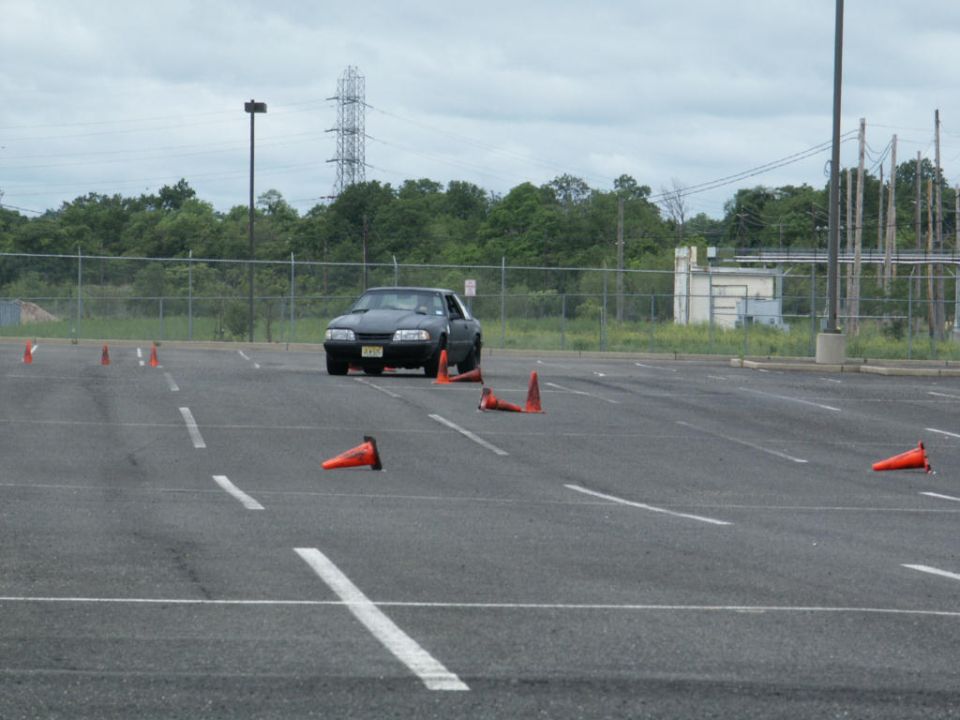 Black coupe with a license plate, angled front view, on a paved lot with orange cones
