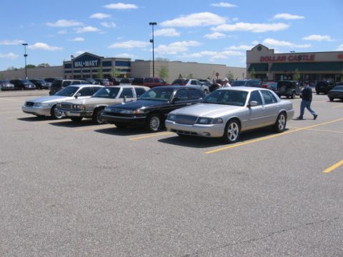 1988 Mercury Grand Marquis - A row of sedans are parked in a lot on a sunny day, with retail stores in the background.