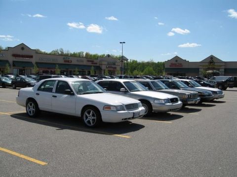 1988 Mercury Grand Marquis - Several sedans are parked in a lot in front of a strip mall under a blue sky with scattered clouds.