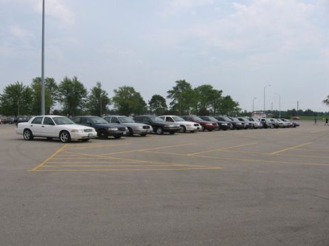 1988 Mercury Grand Marquis - A line of sedans, mostly dark colored with a few white ones, are parked in an outdoor lot under a cloudy sky.