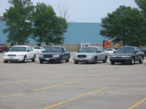 1988 Mercury Grand Marquis - Several sedans are parked in a lot, with a blue building and trees in the background.