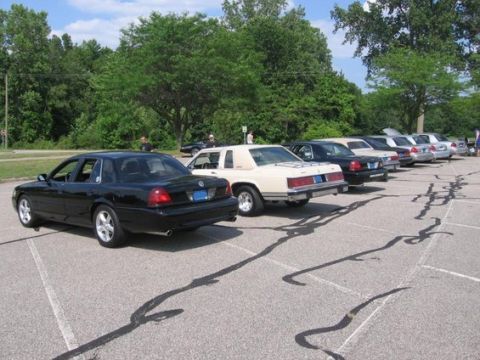 1988 Mercury Grand Marquis - Several sedans are parked in a lot under a cloudy sky, with trees in the background.