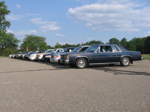 1988 Mercury Grand Marquis - A lineup of sedans, including a dark blue 1988 Mercury Grand Marquis, sits parked under a partly cloudy sky.