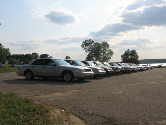 row of silver sedans parked in a lot, some trees and a lake in the background