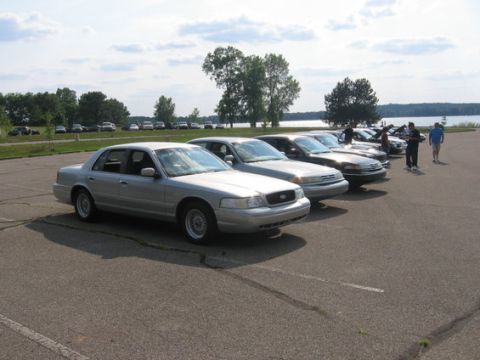 1988 Mercury Grand Marquis - A line of sedans are parked in a lot near a body of water, with several people standing nearby.