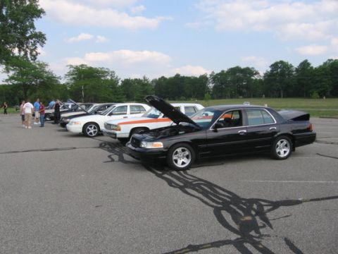 1988 Mercury Grand Marquis - Several sedans are parked in a line on asphalt, with trees and a cloudy sky in the background.