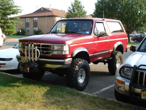 1994 Ford Bronco - Red lifted Ford Bronco parked at the end of an apartment-complex lot, shot from the front three-quarter driver side. It has a winch mounted…