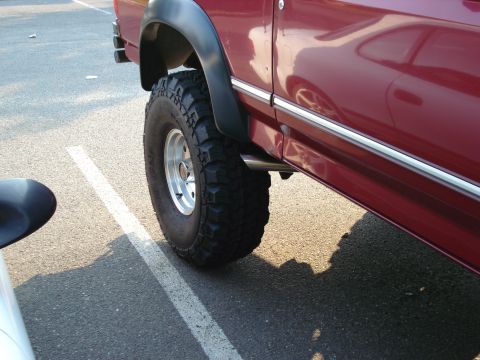 1994 Ford Bronco - Low-angle detail of the front driver-side wheel and rocker area on a lifted red Ford truck in a parking lot, showing a Dirt Grip mud-terrai…