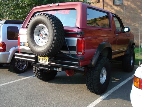 1994 Ford Bronco - Rear three-quarter view of a lifted red Ford Bronco on chrome wheels and mud-terrain tires, with a swing-out tire carrier holding a full-si…