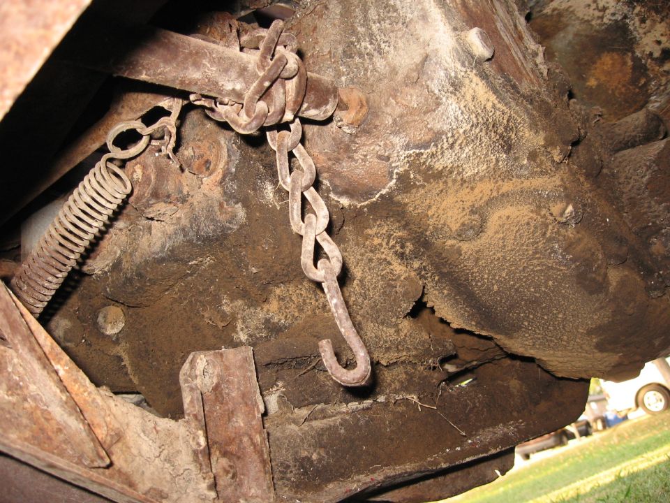 Undercarriage of a rusty vehicle showing a chain and spring mechanism. Some wood bracing is visible