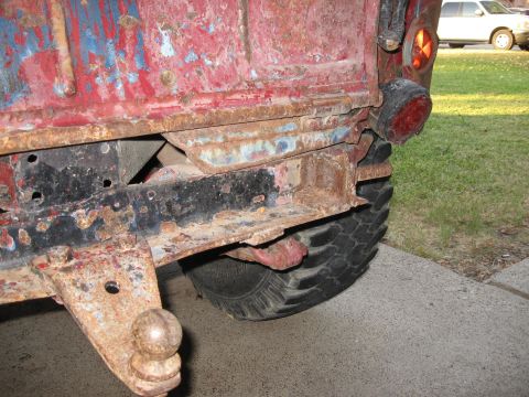 1983 Chevrolet Silverado 1500 - Rear view of a very rusty truck, showing the frame, tow hitch, and tire. Another vehicle is in the background.