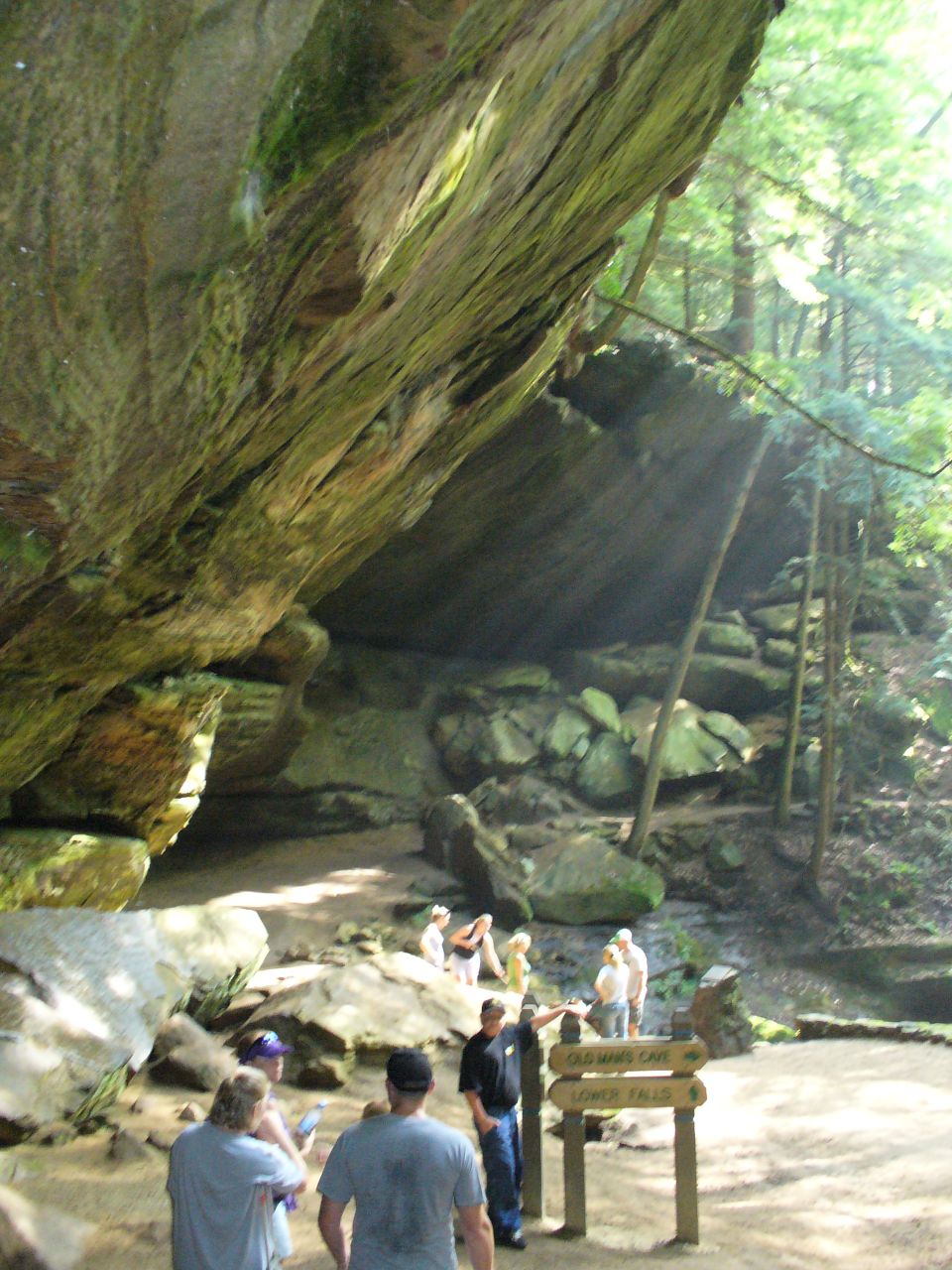 People gathered near a large rock overhang in a wooded area with a signpost