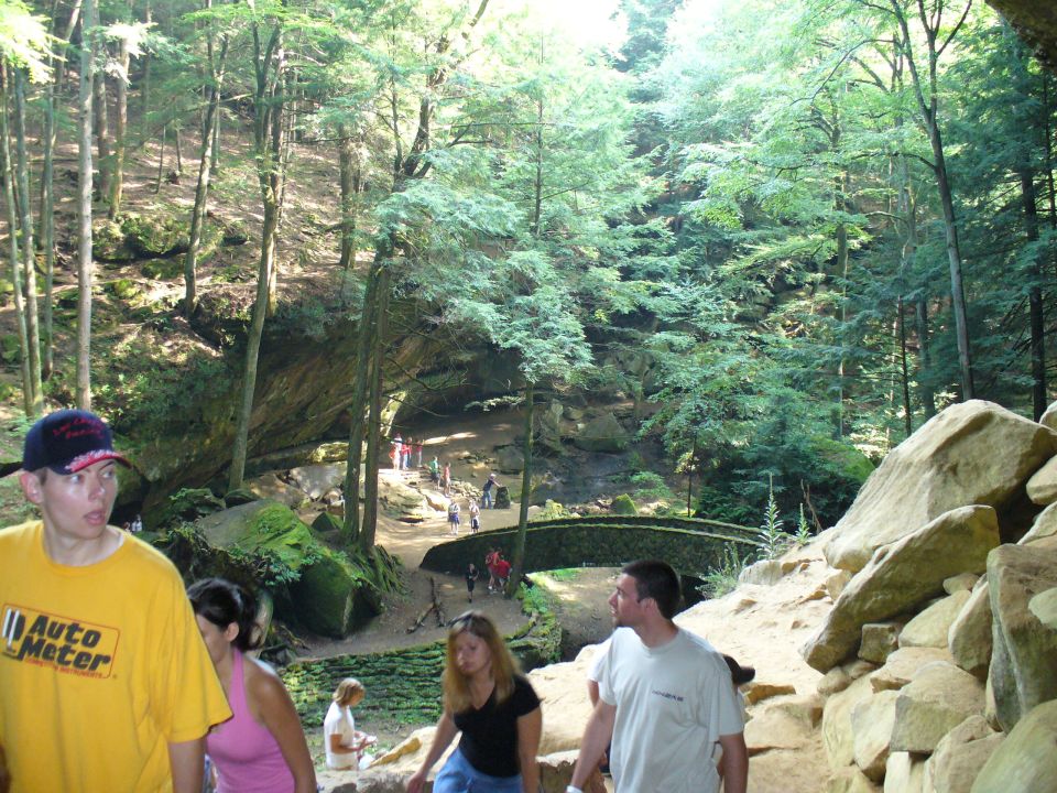 People walk near a stone bridge in a forest with large rock formations and trees
