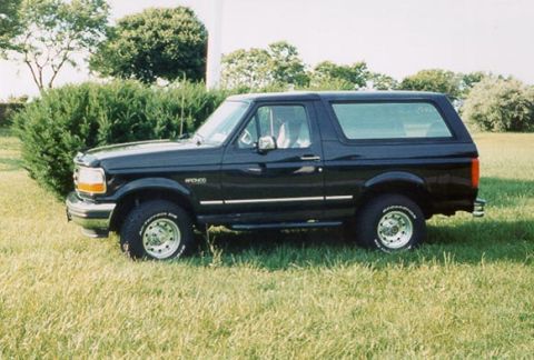 1996 Ford Bronco - A black Ford Bronco is shown from the side, parked in a grassy area with trees in the background.