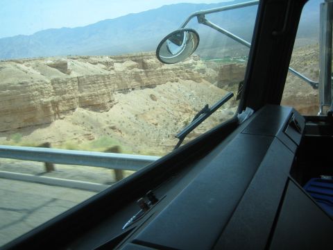 1990 Ford Bronco - Looking out a truck windshield and side mirror at an eroded desert canyon and highway guardrail under hazy skies.