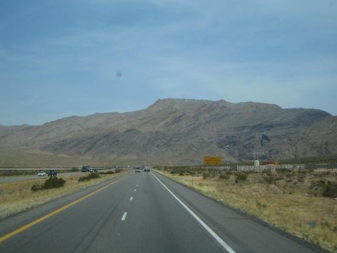 1990 Ford Bronco - multi-lane desert highway stretches toward a rugged tan mountain ridge under a pale blue sky.